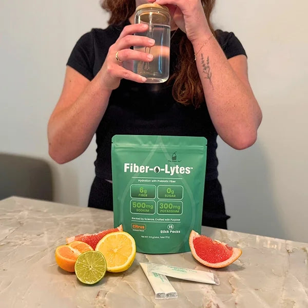 Person drinking from a glass jar beside Fiber-O-Lytes citrus electrolyte mix with fiber, stick packs and citrus fruit