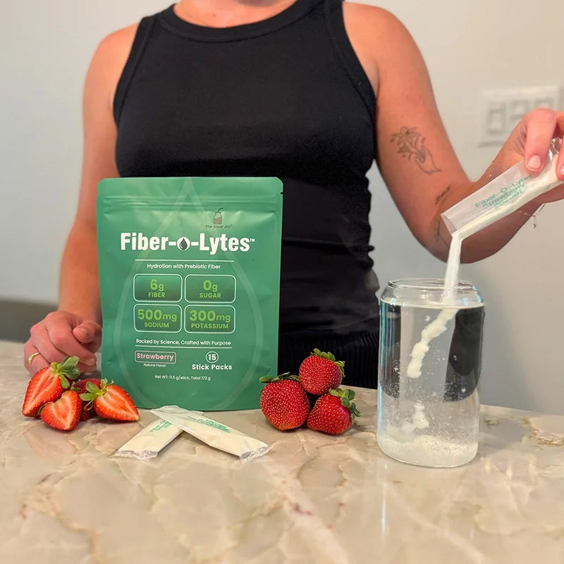 Person pouring Fiber-O-Lytes strawberry electrolyte mix with fiber into water beside the pouch, stick packs and strawberries