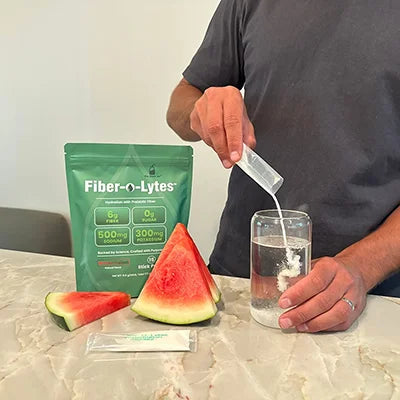 Person pouring Fiber-O-Lytes watermelon electrolyte mix with fiber into a glass of water beside the pouch and watermelon slices